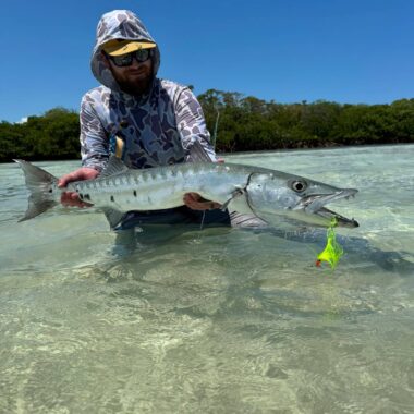 Man wearing a camo hoodie and yellow cap in shallow clear water, holding a large barracuda fish