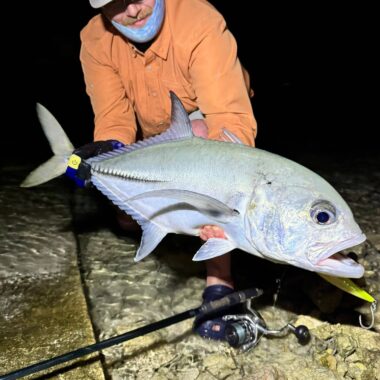 Fisherman in an orange shirt and headlamp holding a large silver fish on a rocky night shoreline, with fishing rod nearby.
