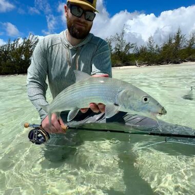Man wearing sunglasses and a beige cap holds a large silver fish in knee-deep, clear turquoise water with mangroves in the background.