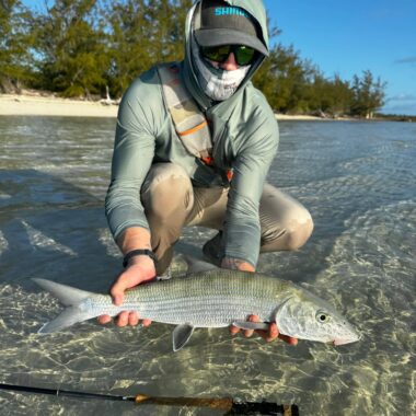 Fisherman kneels in shallow, clear water, holding a large silver fish with a fishing rod nearby and trees in the background.